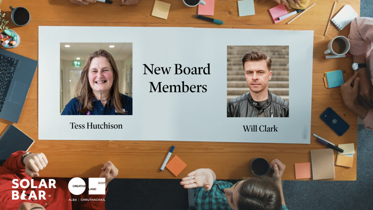 Image - A poster announcing new board members for an organization. The top section reads “New Board Members” in large black text. Below, there are two rectangular photos: the first shows a person wearing a dark blue collared shirt with long hair, labeled “Tess Hutchison.” The second photo shows a person wearing a black leather jacket over a dark shirt, labeled “Will Clark.” The background of the poster is light gray, and the bottom left corner features the Solar Bear logo along with the Creative Scotland logo. Around the poster, there is a wooden table with colorful sticky notes, pens, and coffee cups visible.